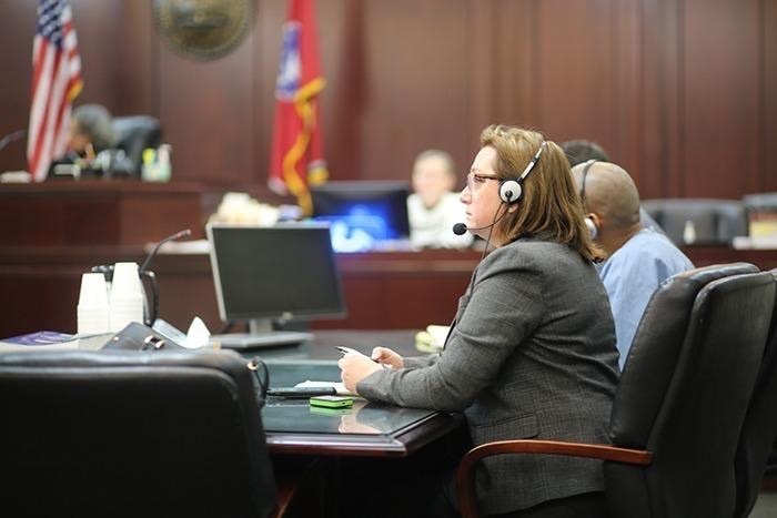 An interpreter wearing a headset listening at a table in a courtroom, the bench and flags behind her, illustrating court and legal interpretation.